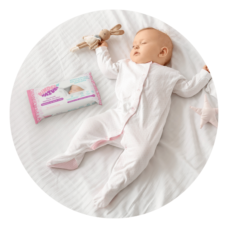 Niemowlę w białej koszulce śpi na białym łóżeczku, trzymając dzianinowego króliczka. Nieopodal, chusteczki nawilżane dla dzieci Kinder by Nature z różowo-niebieskim wzorem spoczywają obok miękkiej zabawki w kształcie gwiazdy w pobliżu wyciągniętej lewej ręki dziecka.