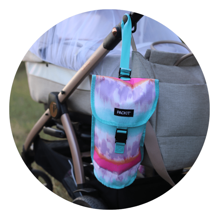 Kolorowa izolowana torba na butelkę z wzorem tie-dye w odcieniach turkusu, różu i fioletu, z napisem "PACKIT", wisi na rączce wózka. Wózek i szara torba na pieluchy są widoczne w tle, na zewnątrz, na trawie.