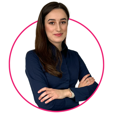 Zespół 13 Kobieta z długimi brązowymi włosami ubrana w ciemnoniebieską koszulę zapinaną na guziki stoi ze skrzyżowanymi ramionami, uśmiechając się delikatnie. Jest otoczona różowym okręgiem na białym tle.