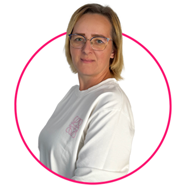 Zespół 45 Kobieta o krótkich blond włosach i w okularach ma na sobie białą koszulę z różowym logo. Stoi bokiem, ale twarzą do kamery, uśmiechając się delikatnie. Tło jest białe, a jej górną część ciała otacza różowy okrąg.